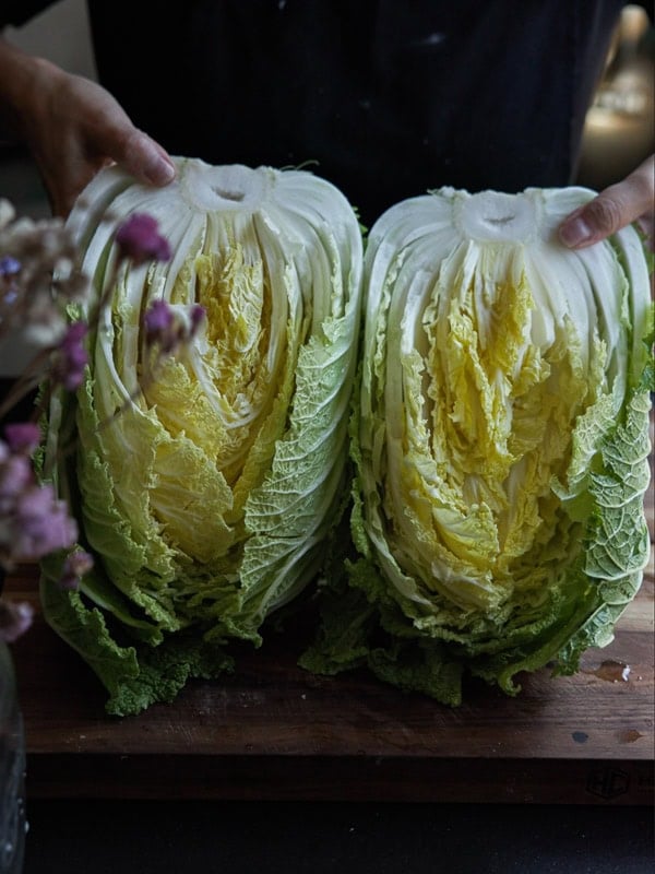 A person holding two halves of a napa cabbage on a wooden cutting board. The cabbage is fresh, with green and yellow leaves, and is surrounded by a few small, out-of-focus pink flowers.